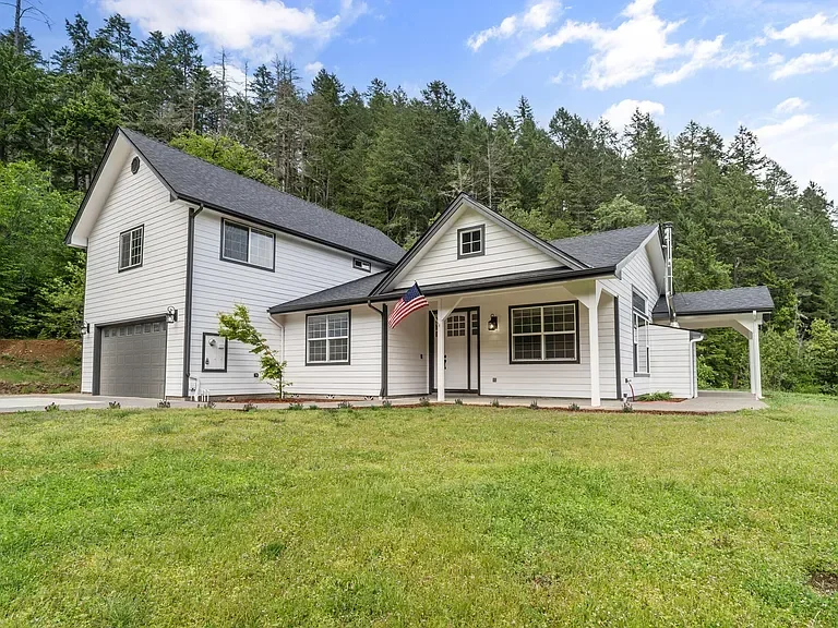 White farmhouse with charcoal roof, covered porch, and attached garage surrounded by evergreen trees